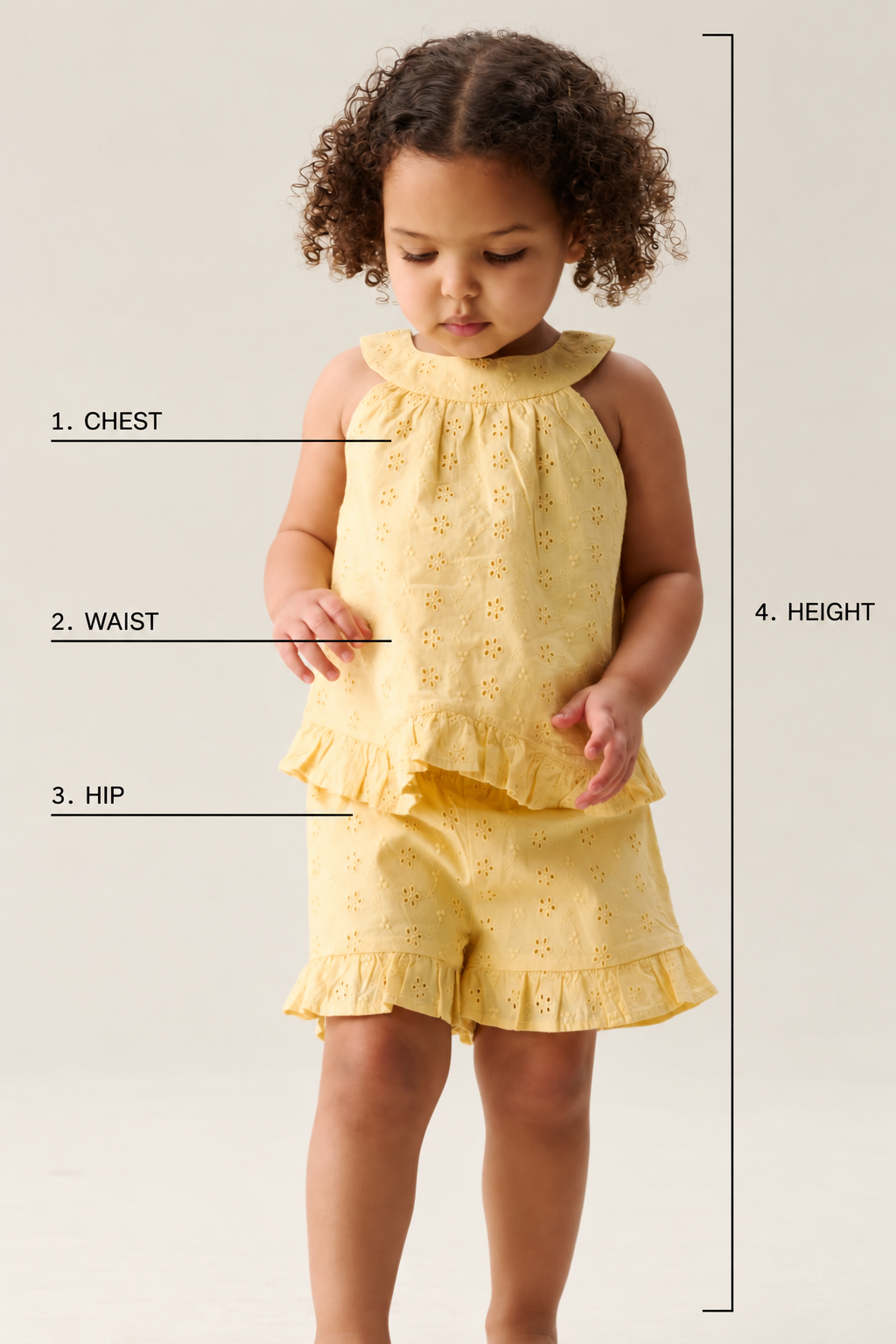 A toddler wearing a yellow eyelet two-piece outfit with a ruffled halter top and matching shorts, shown against a light neutral background with measurement lines and labels indicating chest, waist, hip, and height.